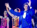 Una escena teatral con un hombre en traje azul brillante y corbata, junto a dos bailarinas en vestidos coloridos.