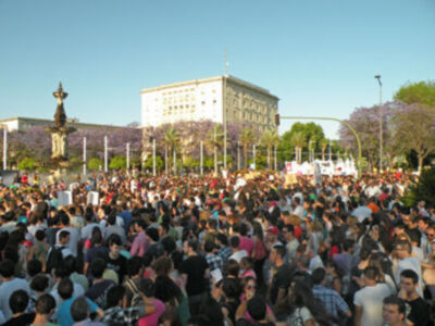 Una multitud en una plaza con árboles y edificios al fondo.