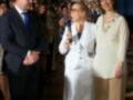 Una mujer con un vestido blanco y una corbata azul, posa junto a otra mujer en un evento formal. La escena parece ser una celebración o reunión importante, con personas vestidas de manera elegante.