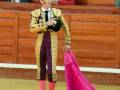 Un torero en traje tradicional de color dorado y morado, con un pañuelo rosa en la mano izquierda. El fondo muestra una arena de toros con asientos y personas observando el evento.