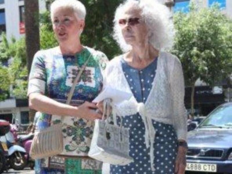 Dos mujeres caminan por la calle, una con un vestido floral y la otra con un vestido azul a lunares, ambos con sombreros de ala ancha. En el fondo, se observa un coche y una motocicleta estacionada.