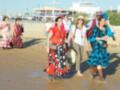 Una multitud de personas vestidas con trajes típicos flamencos caminan por la playa. La imagen muestra a varias personas con vestidos coloridos, botas altas y sombreros tradicionales. La playa está desierta con arena clara, y en el fondo se pueden ver edificios blancos con azulejos. La gente parece estar disfrutando de un día soleado y festivo en la playa.