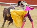 Un torero en acción durante una corrida de toros en España, con el toro en el fondo y el torero con su muleta y capote en el frente.