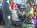 Una mujer en un vestido de colores vibrantes baila en el centro de una reunión festiva, rodeada de personas en trajes tradicionales y sombreros, en un ambiente festivo y colorido.