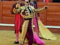 Un torero en traje tradicional de color rojo y dorado, con un capote amarillo, realiza una coreografía en el ruedo de la plaza de toros. El fondo muestra a otros espectadores y toreros en el interior del coso.