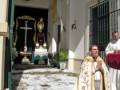 Un sacerdote en un vestido de color dorado y blanco, con una cruz dorada en la parte superior, se encuentra en el centro de la imagen. A su izquierda, hay una mesa con flores y un altar, mientras que a la derecha se puede ver a otro sacerdote con una saya blanca y un manto rojo. La imagen se encuentra en un patio con baldosas blancas, una escalera y plantas verdes.