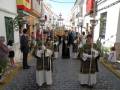 Una procesión religiosa en una calle de la ciudad, con personas vestidas con trajes tradicionales y portando estandartes. El cielo es claro y la arquitectura local se puede ver en el fondo.
