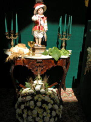 Una escena festiva con un joven en traje colonial sentado en una mesa de madera tallada. La mesa está adornada con flores blancas y un ramo de hojas verdes, junto a velas azules en candelabros. La pared detrás es verde y el suelo tiene un patrón de cuadros.