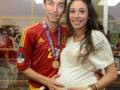Un futbolista con una camiseta de España y medalla dorada posa junto a una mujer embarazada, ambas sonrientes. El fondo muestra un ambiente festivo con carteles y luces.