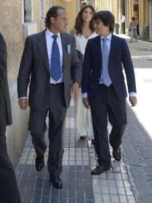 Dos hombres en trajes de negocios caminan por una calle. La imagen muestra a dos personas vestidas formalmente, uno con un traje azul y corbata azul, y la otra con un traje negro. La mujer a su lado lleva un vestido blanco largo y zapatos de tacón. En el fondo, se pueden ver edificios y una calle con gente caminando.