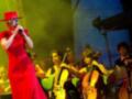 Una mujer en un vestido rojo largo y sombrero rojo canta mientras un grupo de músicos toca instrumentos en una escena de concierto.
