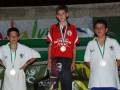Tres niños en un podio, dos con medallas y una bandera de fondo.