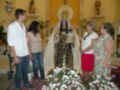 Una imagen de una Virgen María vestida en traje negro y dorado, con un manto blanco, rodeada de flores blancas y rosadas. En el fondo, una estatua de la Virgen en un altar con velas encendidas y adornos religiosos. A su alrededor, cuatro personas observan la imagen con atención y respeto.