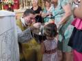 Una imagen de una niña en un vestido blanco con lunares, observando algo interesante mientras una mujer toma su foto. La escena parece ser en un ambiente religioso, posiblemente una iglesia, con detalles como velas y adornos tradicionales.