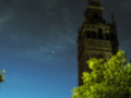 La Giralda, la torre de la Catedral de Sevilla, sobresale en el cielo nocturno con su cúpula dorada y detalles en piedra. La luna y un planeta se alinean con la estructura, creando una escena celestial impresionante.