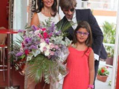 Una mujer sonríe mientras sostiene un gran ramo de flores, acompañada por una niña en un vestido coral y un hombre con gafas de sol. En el fondo, se observa una terraza con plantas y una mesa roja.