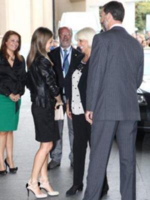 Una mujer con vestido negro y zapatos de tacón blanco se dirige a saludar a una persona en traje. Detrás, dos hombres y una mujer con vestido negro miran a la persona en traje.