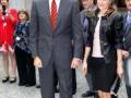 El rey Felipe VI y la reina Letizia de España caminan juntos en una ceremonia formal, ambos con trajes elegantes. La reina lleva un vestido negro y sandalias de tacón, mientras que el rey usa un traje gris con corbata roja. La imagen captura una escena de protocolo real en un evento importante.