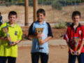 Tres niños sonríen, cada uno sostiene un trofeo. El niño en el medio lleva una camiseta azul con un trofeo dorado. El niño a la izquierda lleva una camiseta amarilla con un trofeo azul. El niño a la derecha lleva una camiseta roja con un trofeo dorado. En el fondo, hay un parque y vehículos aparcados.