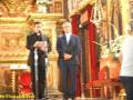 Imagen de una iglesia con dos hombres en trajes formales, uno frente a un altar dorado y otro de espaldas. Hay una persona en la derecha, parcialmente visible, con un vestido blanco y cabello largo. La decoración dorada y roja del interior sugiere una ceremonia religiosa o formal.
