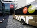Un bus de Axor, una empresa de transporte, estacionado frente a un edificio de oficinas moderno con su logotipo visible en la parte trasera del vehículo.