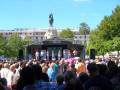 Una multitud en una plaza pública, con un escenario en el centro donde se encuentra una estatua de un rey. El público está sentado y algunos están de pie, observando la escena. La estatua es el centro de atención, con un cielo azul claro y árboles verdes alrededor.