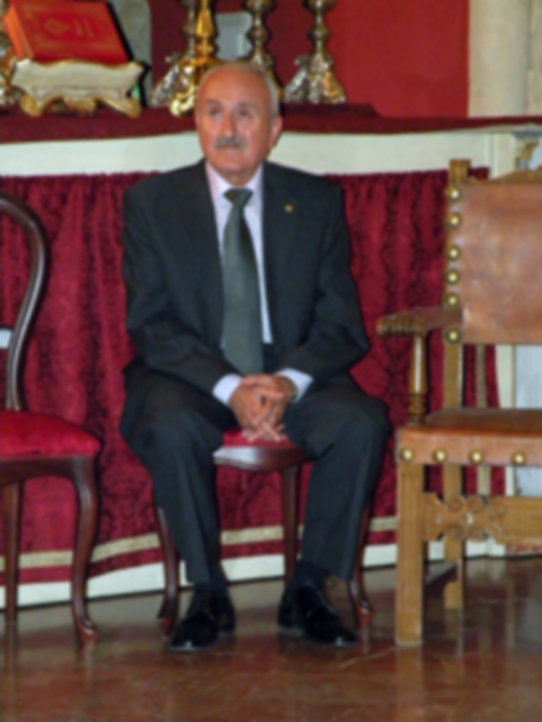 Un hombre en traje negro sentado en un sillón de madera frente a una pared roja con un tapiz. La imagen parece ser de un evento formal o ceremonial.