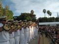 Un grupo de marineros en uniforme blanco, con insignias doradas y sombreros blancos, tocan instrumentos militares en una ceremonia. El cielo es azul con algunas nubes y hay palmeras y árboles en el fondo. La imagen está tomada durante un evento militar o ceremonial.
