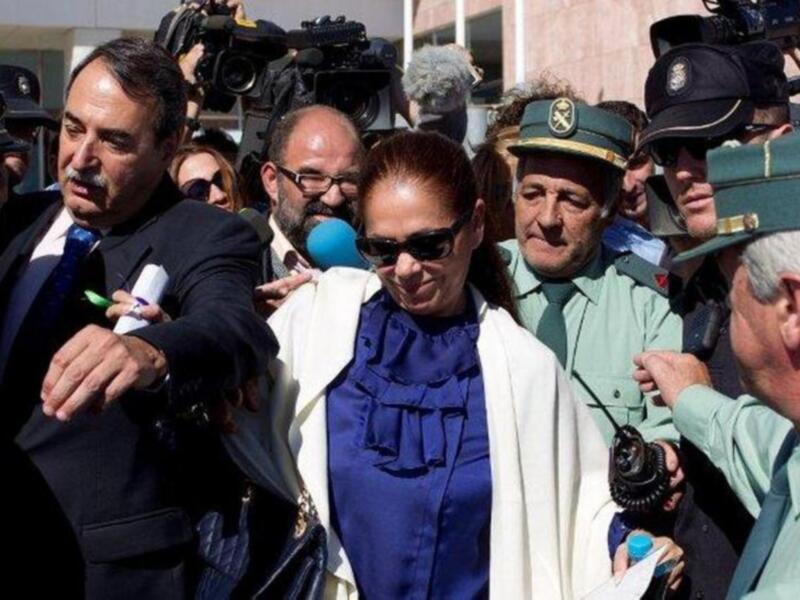 Una mujer con gafas de sol y una blusa azul se dirige hacia la izquierda, rodeada por personas con uniformes militares y medios de comunicación.