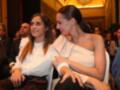 Dos mujeres sonrientes sentadas en un evento formal, rodeadas de otras personas en un ambiente iluminado.