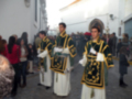 Mujeres vestidas con trajes tradicionales en una calle con edificios blancos y una arquería en el fondo.