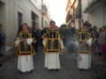 Tres personas vestidas con trajes tradicionales de color negro y dorado, posiblemente en un evento ceremonial o festivo. La imagen muestra una calle con edificios blancos y detalles arquitectónicos, y a la derecha se observa una mujer con un vestido rosa.