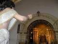 Una escena religiosa dentro de un templo, con una figura vestida en traje dorado y guantes, extendiendo el brazo hacia un altar con una imagen de Cristo. El arco del templo tiene inscripciones y detalles dorados, creando un ambiente de veneración.