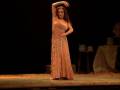 Una mujer en un vestido largo de color naranja está actuando sobre una escena teatral con una mesa y objetos en el fondo.