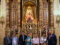 En una iglesia, un grupo de personas posa frente a una imagen religiosa, rodeada por flores y velas. La escena transmite un ambiente festivo y religioso.
