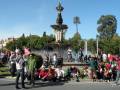 Una multitud de personas disfruta de un día soleado en un parque con una fuente central y árboles verdes alrededor.