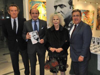 A cuatro personas, tres hombres y una mujer, posando juntos en un evento. La mujer está sosteniendo un libro con la imagen de un hombre en su portada, mientras los hombres están vestidos con trajes formales. En el fondo, hay pinturas y una imagen de un hombre en blanco y negro. La escena parece ser en un museo o galería de arte, con una atmósfera formal y cultural.
