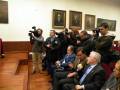 Una sala con paredes blancas, donde se encuentran varios hombres sentados en una mesa. Alrededor de ellos hay personas con cámaras fotográficas, capturando el momento. La escena parece ser una conferencia o reunión formal.