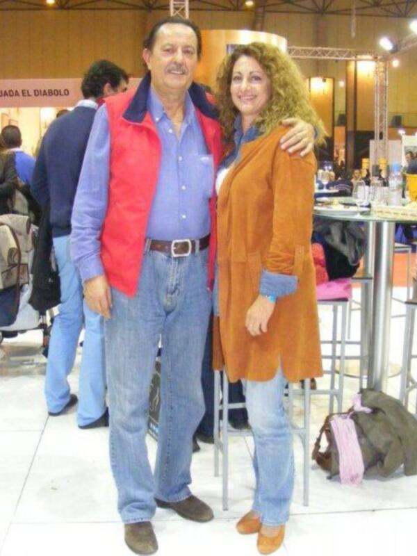 Dos personas posan juntos en un evento. La persona de la izquierda lleva una chaqueta roja y vaqueros, mientras que la persona de la derecha está vestida con un abrigo naranja y vaqueros. Ambos están sonrientes y parecen estar en una feria o evento público, con otros stands y personas al fondo.