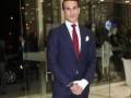 Un hombre en un traje azul con corbata roja y pañuelo de bolsillo blanco. Está en una entrada de edificio con cristales transparentes y luces detrás.