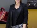 A una mujer con un estilo elegante, posando frente a varios bolsos de lujo. Se puede notar su vestimenta formal y la presencia de varios bolsos de marca en el fondo.