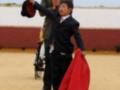 Una escena en un ring de toros, con un niño vestido formalmente sujetando una capa roja mientras un hombre adulto lo guía.