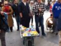 Un perro de raza grande está junto a una carretilla con varios productos en una calle peatonal, con personas caminando alrededor.