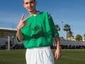 Un joven con una camiseta verde y pantalones blancos posa en un campo de fútbol, con palmeras y una estructura blanca al fondo.