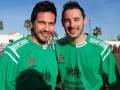 Dos hombres sonrientes con camisetas verdes de fútbol, uno con un logo rojo y otro con una imagen de figuras humanas. Fondo de un campo de fútbol con palmeras y estructuras de apoyo.