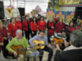 Una agrupación de personas cantando y tocando instrumentos en un ambiente festivo, posiblemente en una escuela o centro comunitario, con decoraciones navideñas en el fondo.