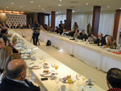 Una reunión de negocios en un salón con mesas largas llenas de platos y copas. Los asistentes están sentados en sillas, conversando y tomando notas. La decoración es formal con cortinas blancas y columnas de madera.