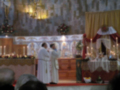 Dos sacerdotes en vestimenta religiosa se encuentran frente a un altar decorado con flores y velas, en una iglesia con mosaicos y vitrales.