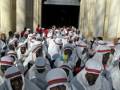 Una procesión religiosa con personas vestidas en trajes tradicionales blancos y rojos, llevando banderas. La imagen muestra una entrada monumental con arcos y columnas blancas, probablemente a un templo o iglesia. La gente parece estar en una celebración o ceremonia importante.