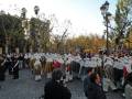 Una procesión religiosa en un parque con personas vestidas de blanco y oro, seguidas por una multitud. El cielo es claro y las hojas de los árboles están caídas, indicando otoño.